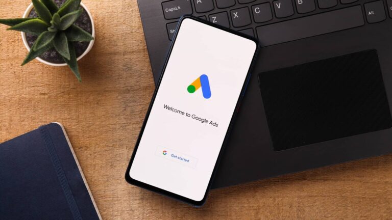 A smartphone on a wooden desk displays the Google Ads welcome screen, next to a laptop keyboard, a notebook, and a small potted plant.
