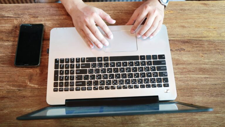 Person using a laptop on a wooden table with a smartphone placed to the left of the laptop.