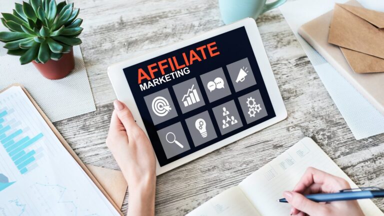 Person holding a tablet displaying "Affiliate Marketing" with related icons, next to a notebook, pen, and a potted plant on a wooden desk.