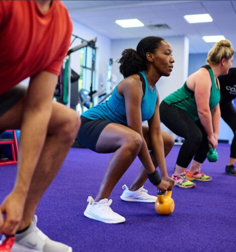 Three people in athletic wear perform squats while holding kettlebells on a purple gym floor.