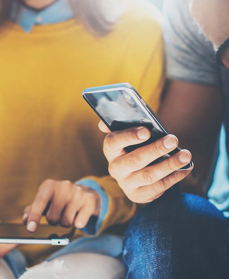 Two people sitting close together, each holding and using a smartphone. The focus is on their hands and phones.