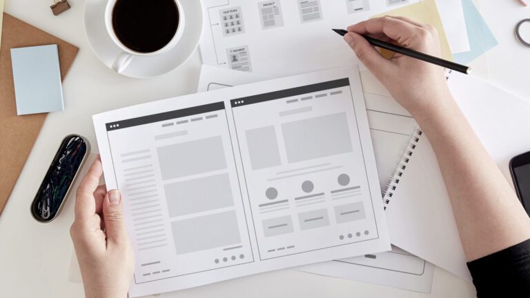 Person holding a website wireframe sketch at a desk with a cup of coffee, a notebook, pen, and various papers and sticky notes.