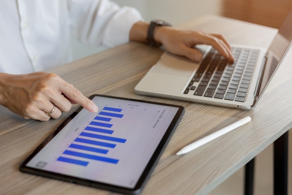 A person points at a bar chart on a tablet while using a laptop on a wooden desk, with a stylus lying nearby.