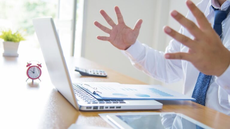 A person in business attire gestures with open hands at a desk with a laptop, charts, a calculator, alarm clock, and a tablet.