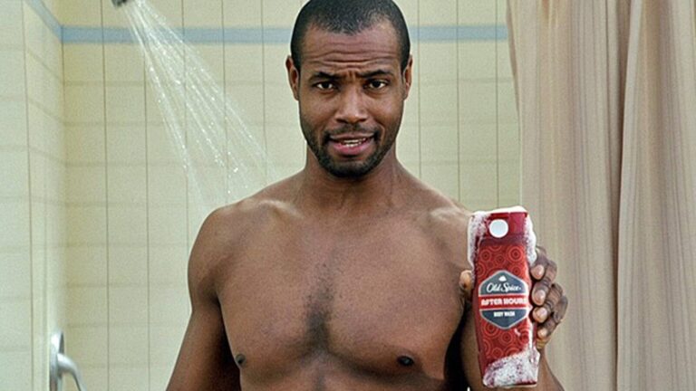 A man stands in a shower holding a bottle of Old Spice body wash, with water running in the background.