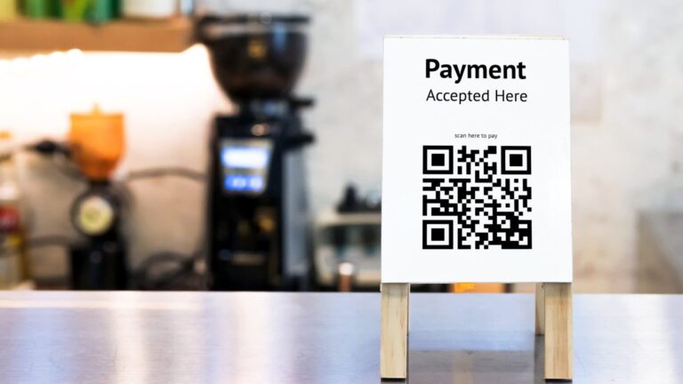 A payment sign with a QR code stands on a counter, indicating "Payment Accepted Here" and prompting users to scan to pay. Coffee shop equipment is blurred in the background.