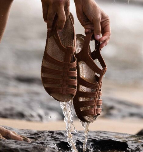 A person holds a pair of brown woven sandals with water dripping from them over rocky ground near a beach.