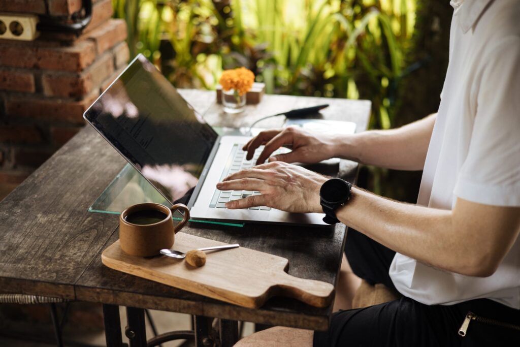 Person typing on a laptop at an outdoor table, perhaps working for a media agency, with a coffee cup, wooden cutting board, and small flower arrangement nearby.