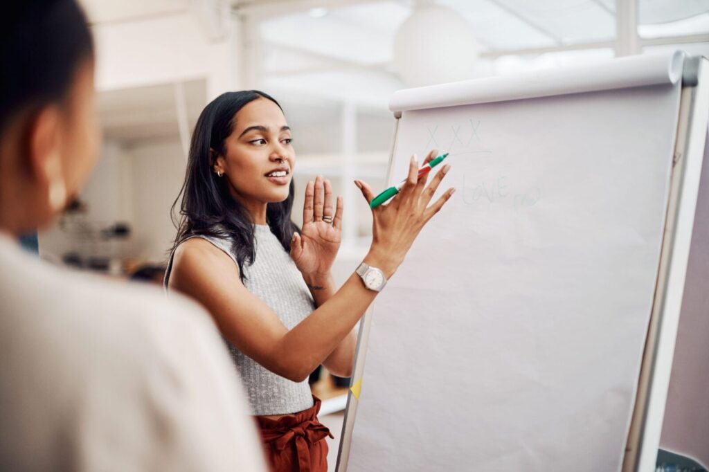 A woman stands by a flip chart, holding a marker and gesturing as she explains something to another person in an office setting.