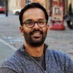 A man with glasses and a beard, wearing a gray sweater, stands outdoors on a city street, smiling at the camera.