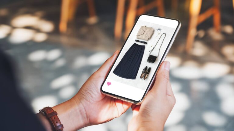 Person holding a smartphone displaying a photo of women's clothing and accessories, including a beige top, black skirt, shoes, bag, and necklace, arranged on a flat surface.