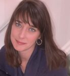 A woman with long brown hair, blue eyes, and hoop earrings looks up at the camera, smiling slightly. She is indoors and wearing a dark top.