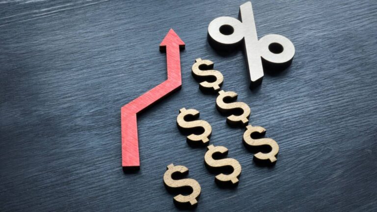 A red upward arrow, several dollar signs, and a percentage symbol arranged on a dark surface, symbolizing rising interest rates or financial growth.